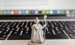 Photo showing a figurine of a wizard, holding a staff, gazing over a Macbook keyboard. In the distance, a document rises up