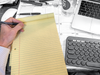 Photo of a hand, beginning to write on a blank legal pad, with computer keyboards at the side.