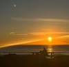 Vivid orange sunset over a calm ocean. In the foreground, a person in silhouette working at a laptop on a picnic bench.