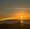 Vivid orange sunset over a calm ocean. In the foreground, a person in silhouette working at a laptop on a picnic bench.