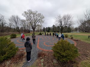Photograph of people walking a labyrinth laid out in brick and gravel in a field, among bare trees, under an overcast sky.